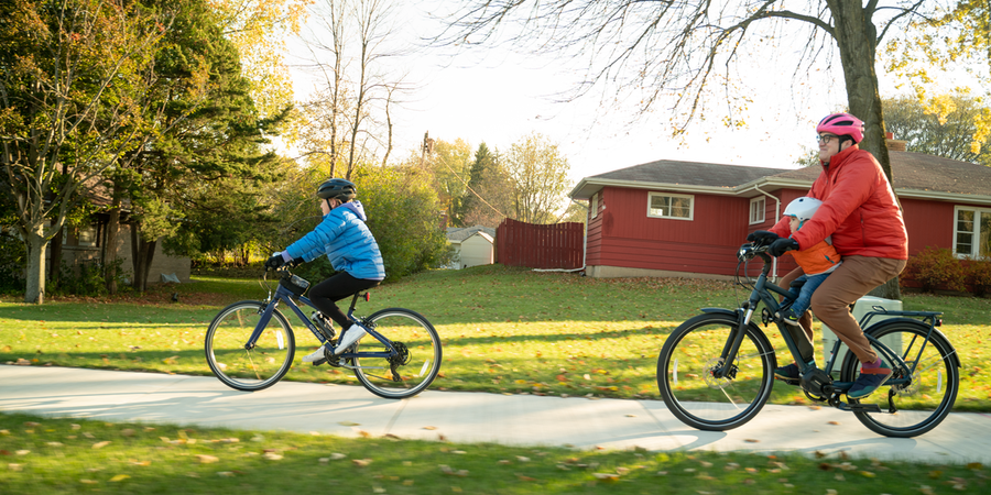 A child bikes in front of an adult on neighborhood sidewalk