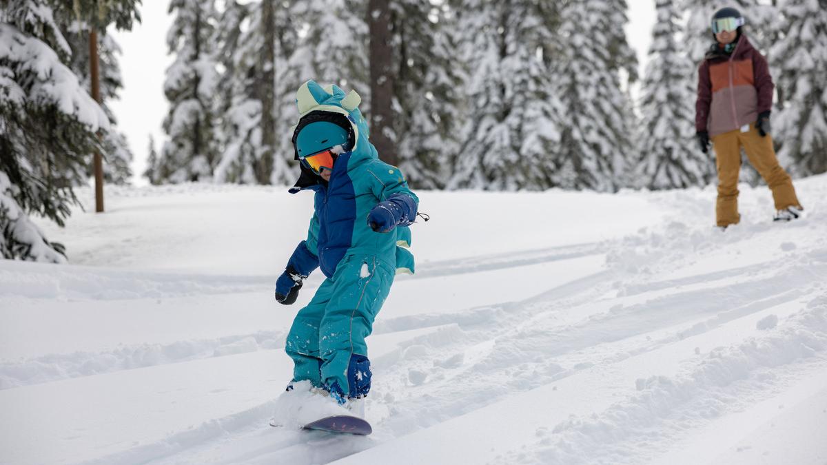 Small child riding a snowboard while an adult looks on in the background