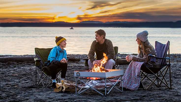 Two adults and a child sitting around fire pit at beach at sunset