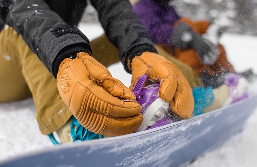 A snowboarder straps into their snowboard while wearing Hestra Fall Line 3-Finger Mittens