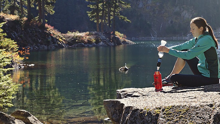 A hiker refills her water bottle from a lake using a squeeze filter