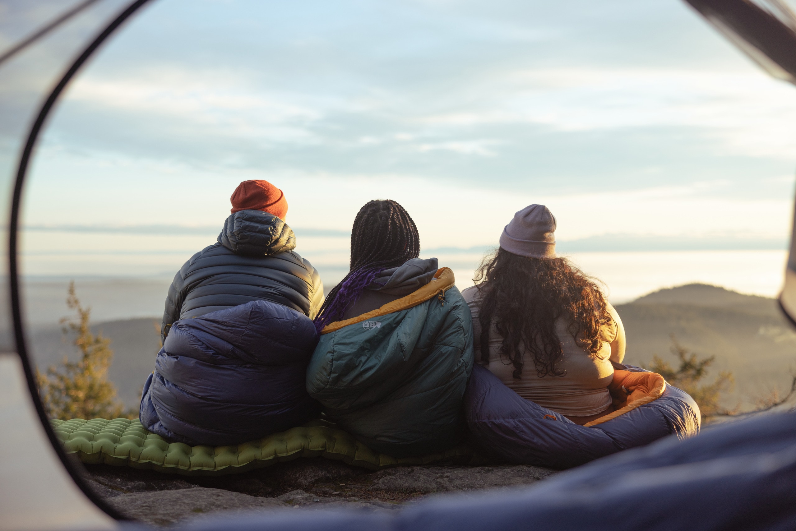 Three backpackers sit in their sleeping bags while looking out at a vista