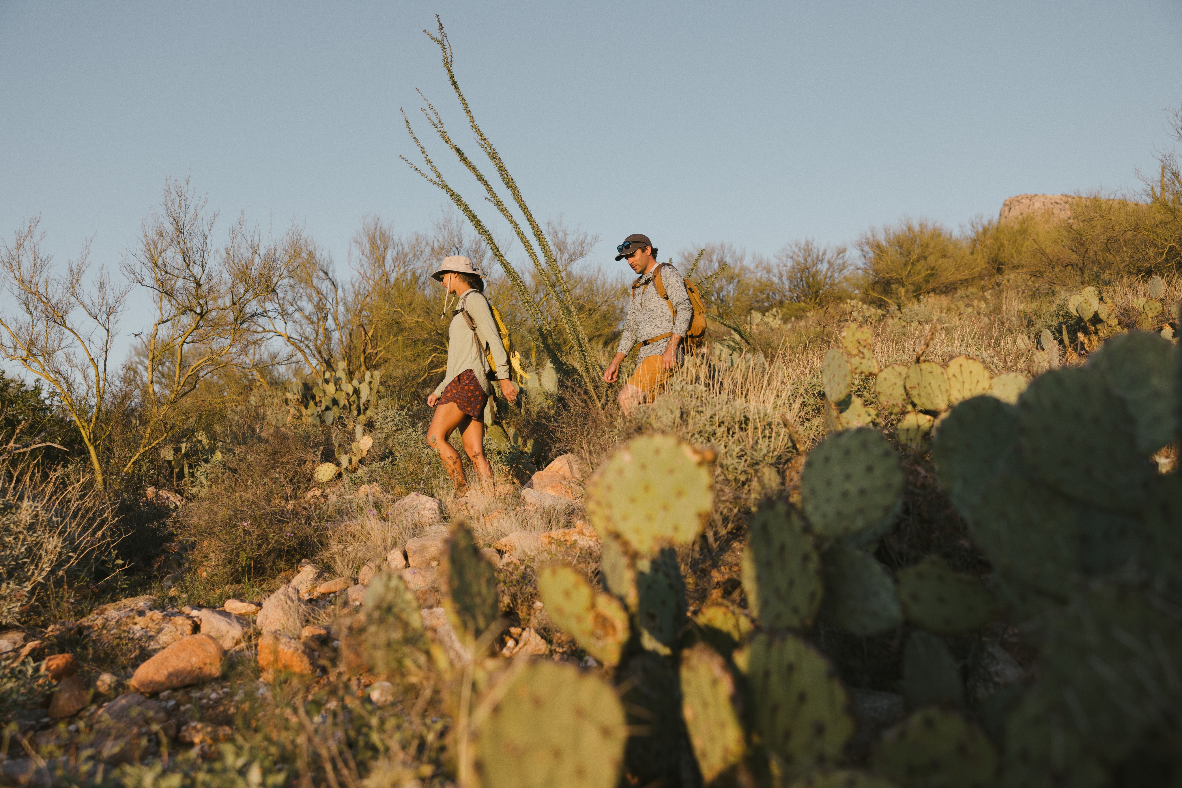Two people hike through a desert trail with large rocks and cactus.