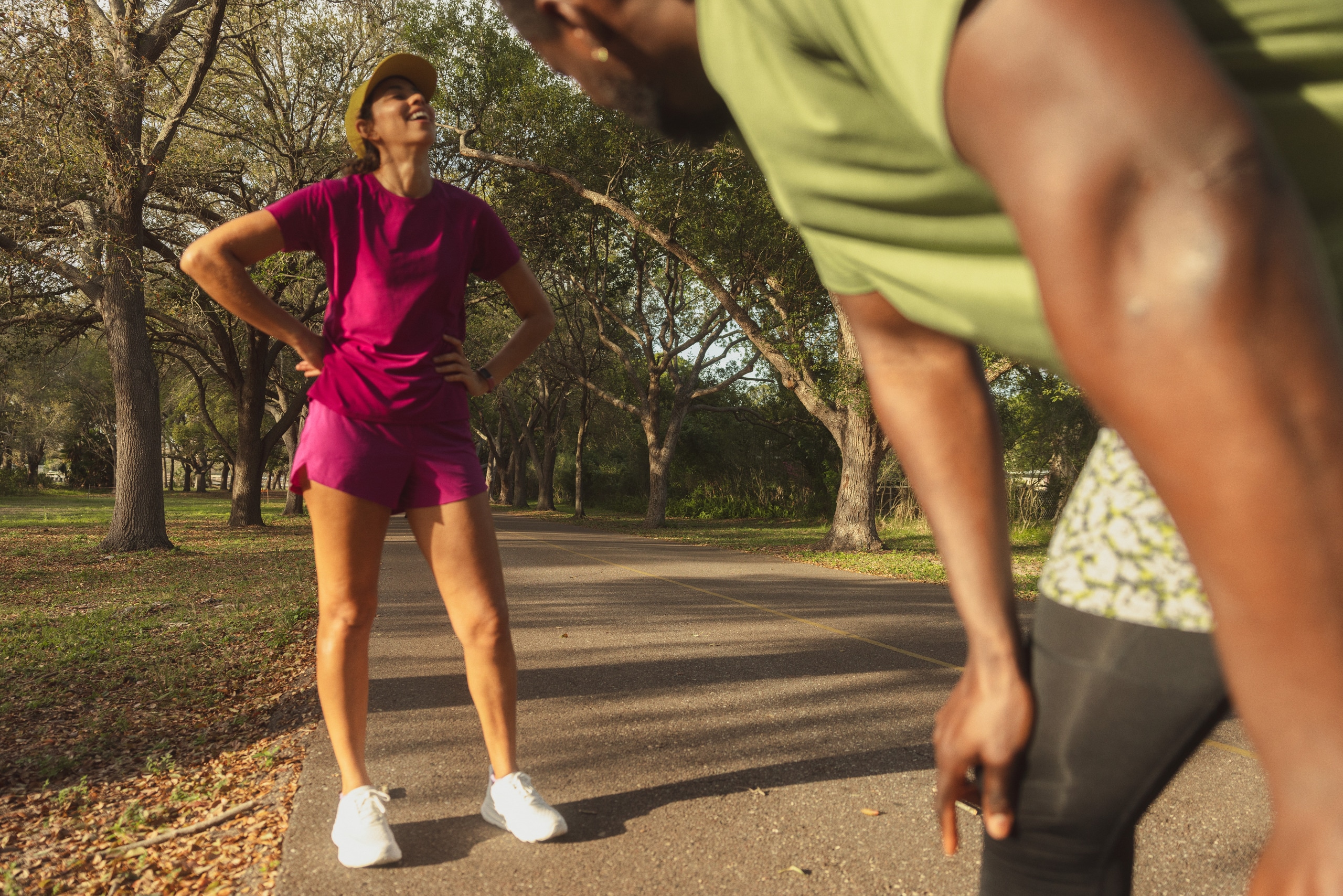 A person in a magenta running outfit and white running shoes smiles, looking up, with their hands on their hips, while another person in green and black running clothes rests with their hands on their knees in the foreground.
