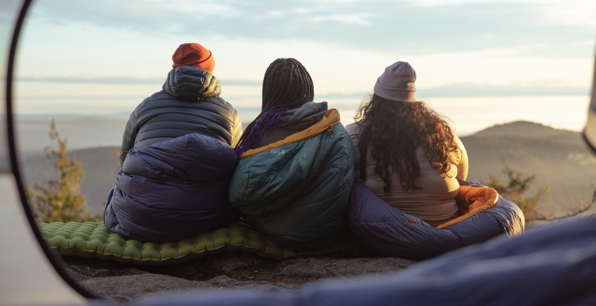 Backpackers bundled up in sleeping bags, looking out over a mountaintop vista.
