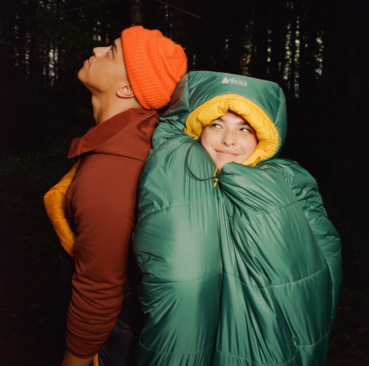 A person smiles, bundled up in a sleeping bag leaning against another person