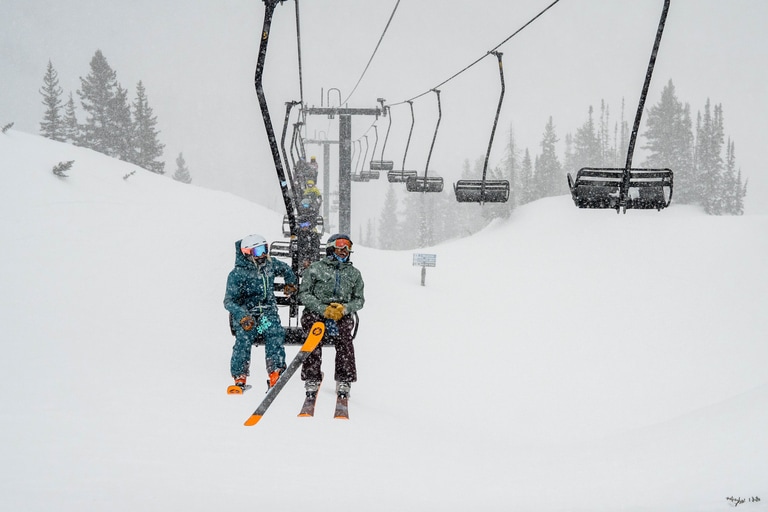 Two skiers using Blizzard Black Pearl skis sit on a chair lift on an overcast day