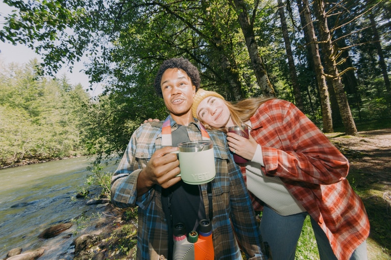Two people hold camp coffee mugs in the woods