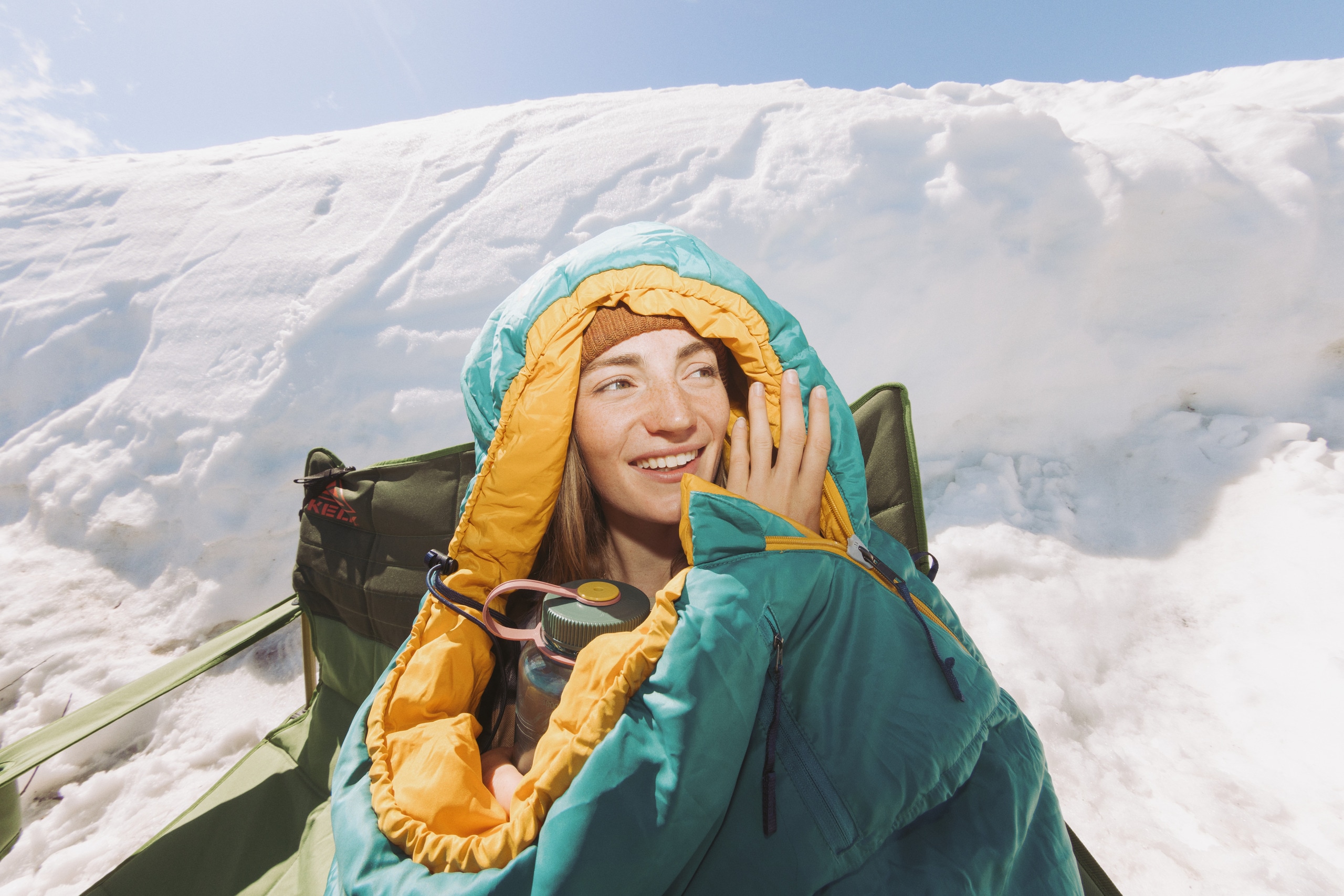 A camper sitting on a camping chair in the snow, wrapped in the REI Co-op Trailmade 20 sleeping bag.