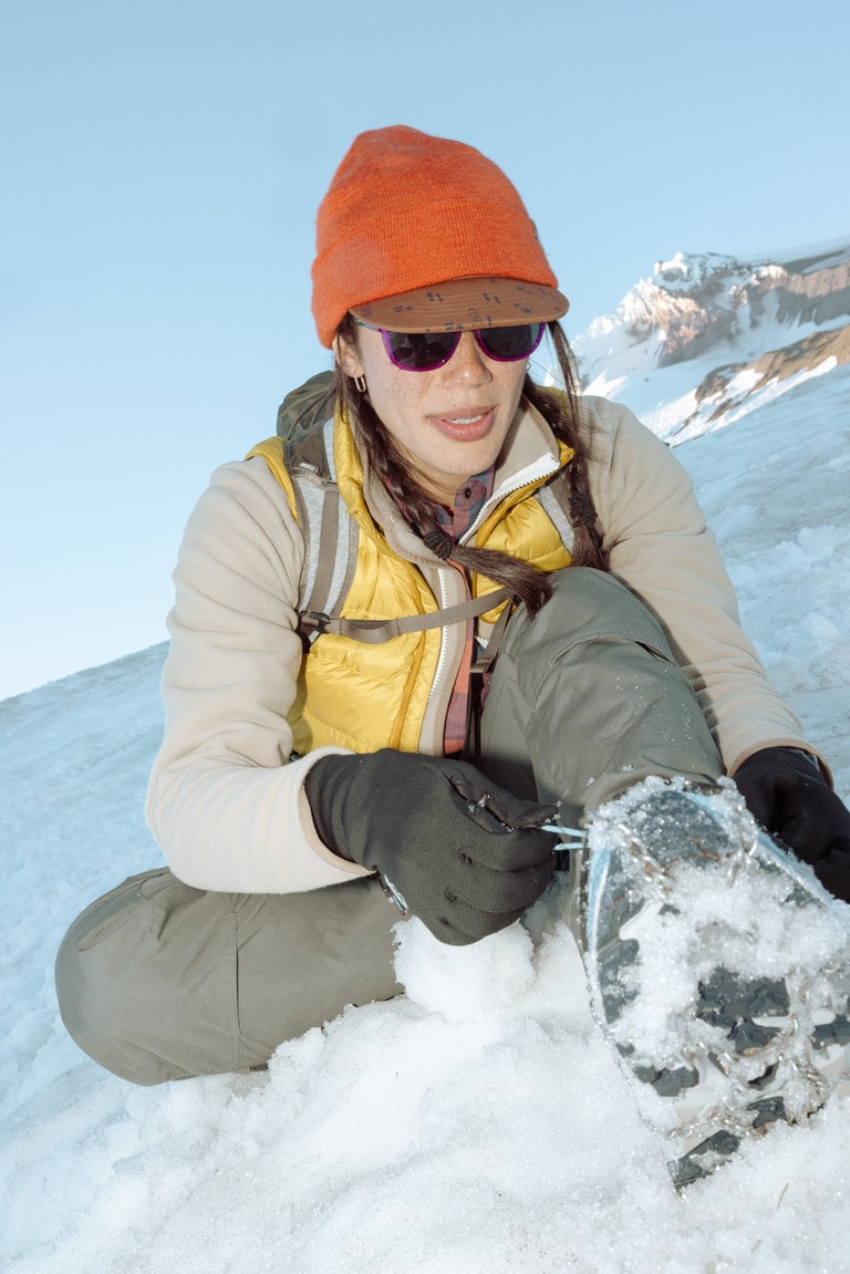 A hiker wearing gloves sits in the snow and ties boot laces.