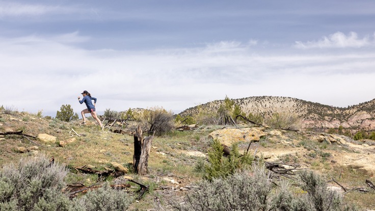 A trail runner crests a sloping plateau with sagebrush and pinon juniper in the foreground.