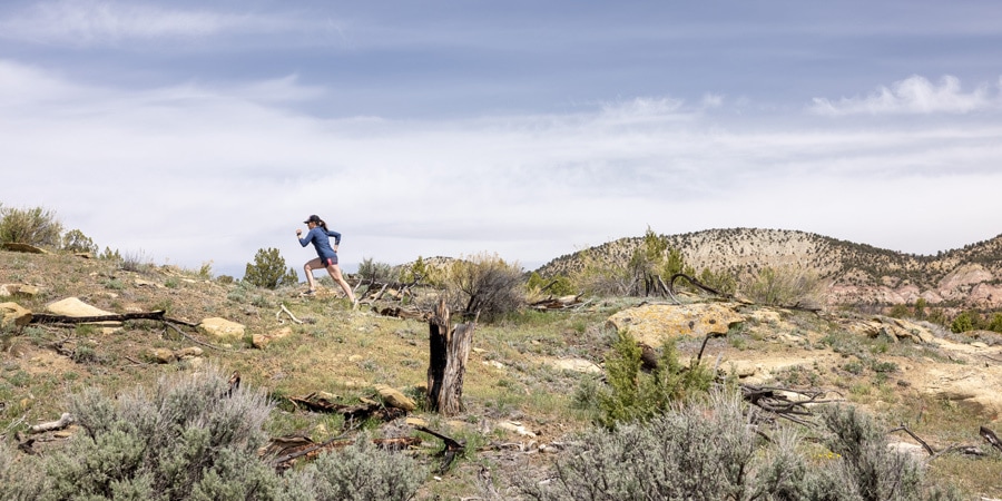 A trail runner crests a sloping plateau with sagebrush and pinon juniper in the foreground.