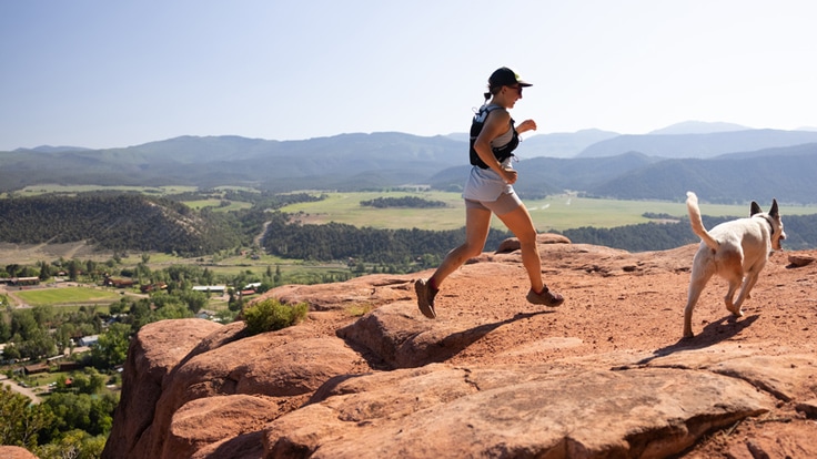 A runner makes her way up a red rock mountain wearing a hydration vest with her dog in tow.