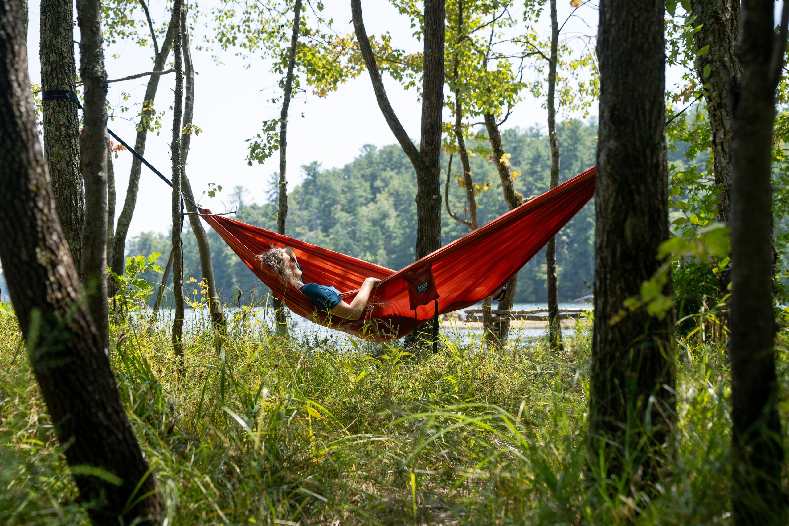 A person lounges in a red ENO DoubleNest hammock between tall trees next to a body of water.