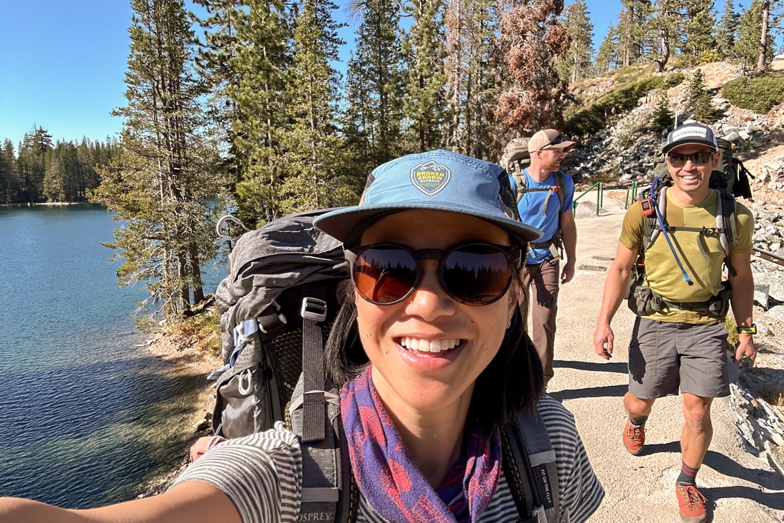 A young woman takes a selfie with her friends in the background while hiking together on a sunny day.