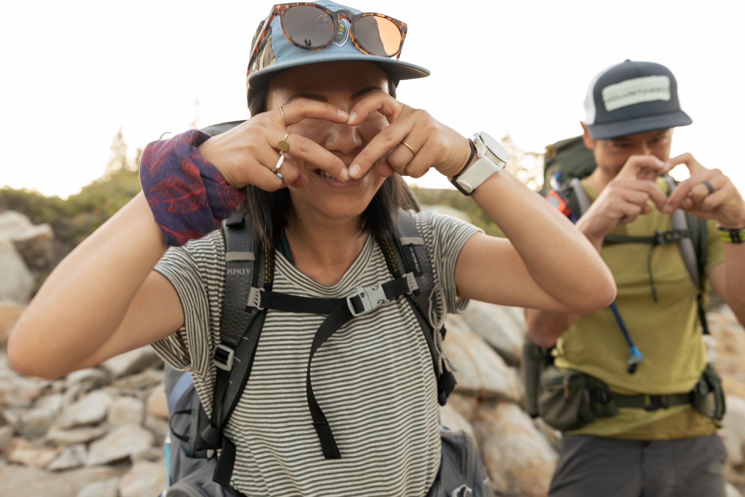Two people wearing hiking gear give finger hearts to the camera