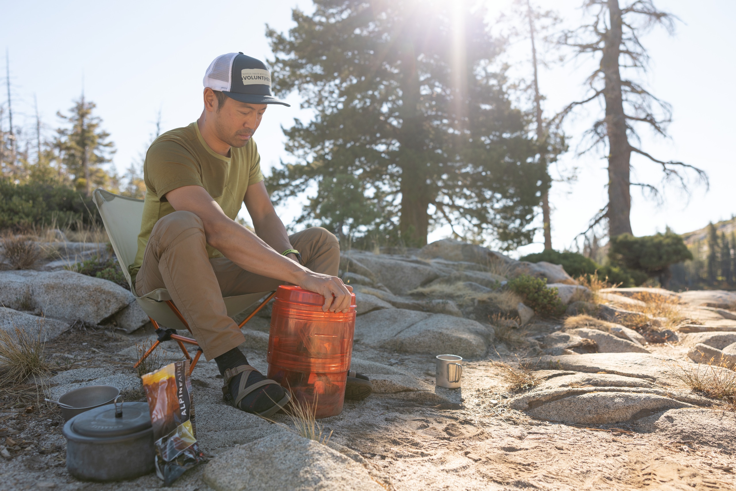 A person sits in a camp chair next to a backpacking stove and is about to open an open bear canister serving food.