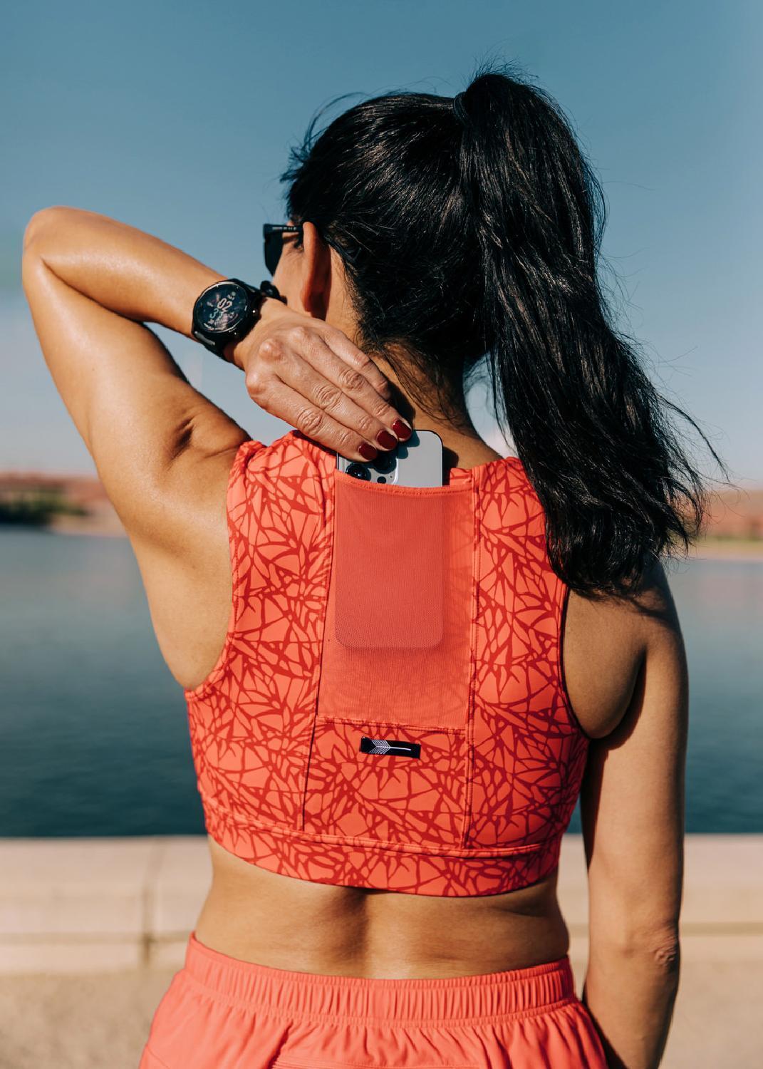 A rear view of a woman in a peach-colored Oiselle Flyout Bra, placing her phone into the bra's mesh-sleeve pocket.