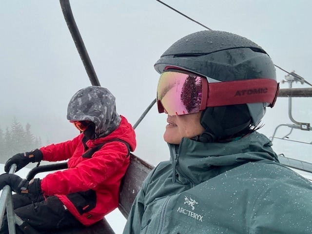 An adult skier and a child snowboarder ride the chairlift, both wearing ski helmets and goggles 