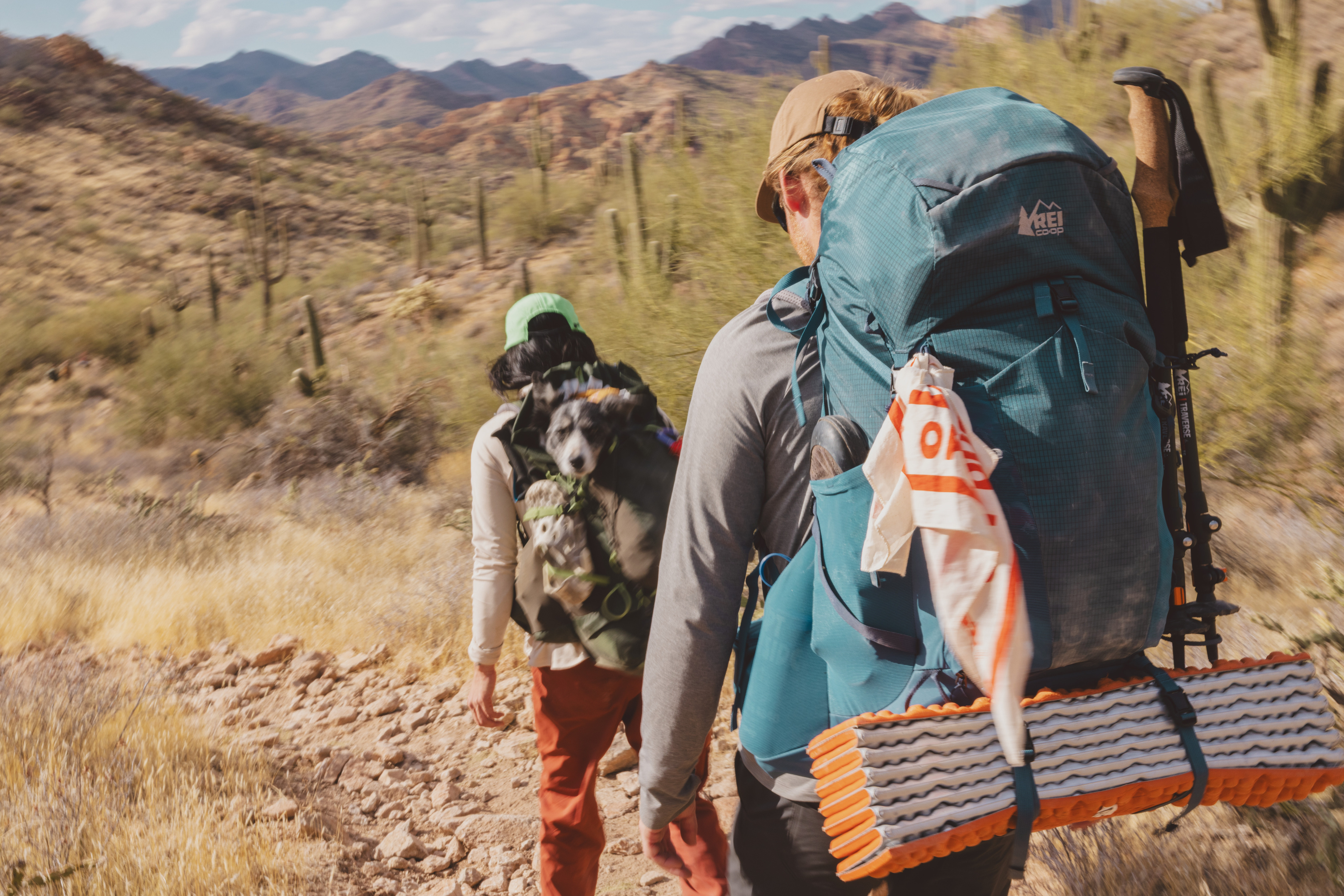 Two backpackers hike through a rocky trail with their dog.