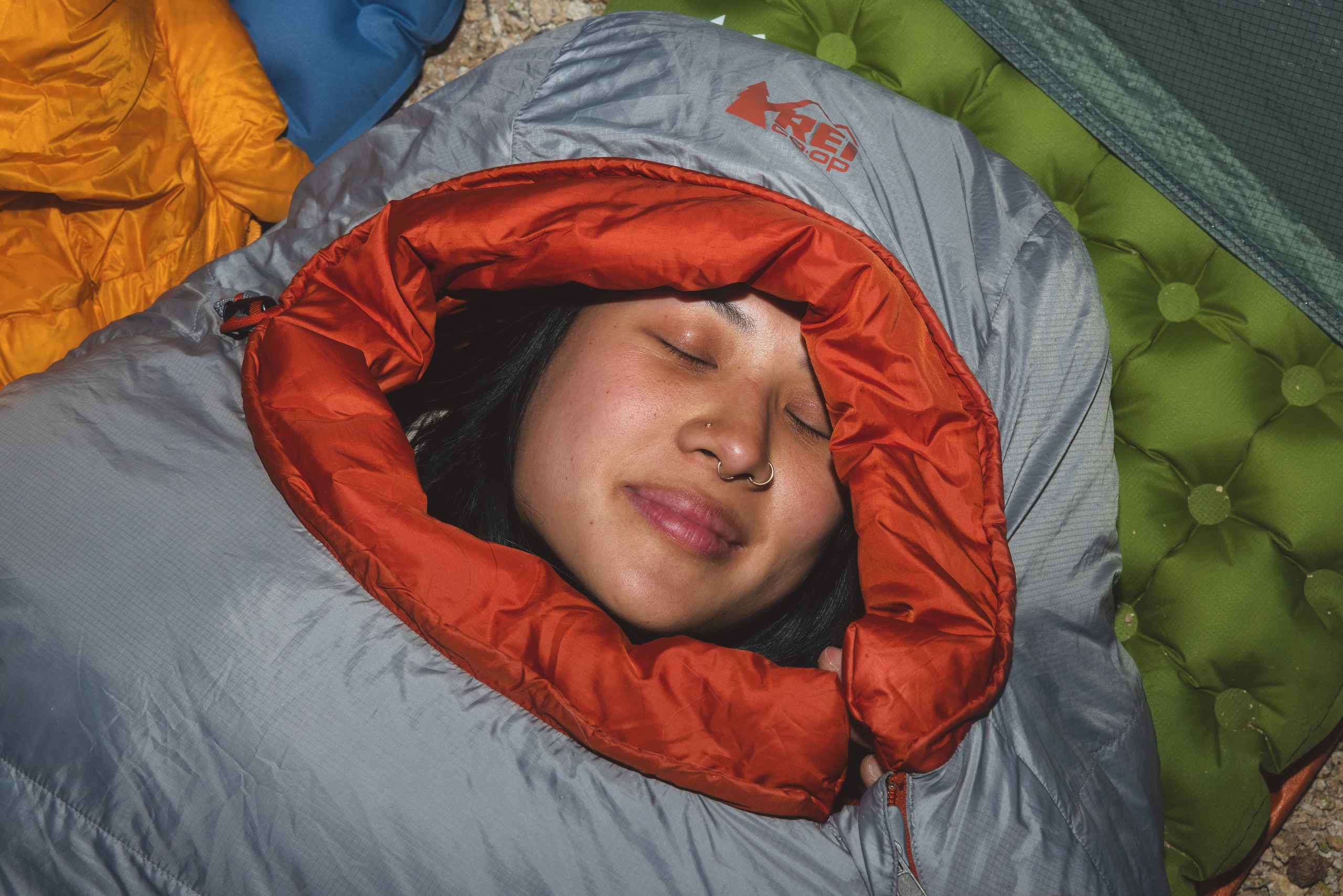 A person smiles with eyes closed while snuggled in a gray sleeping bag 