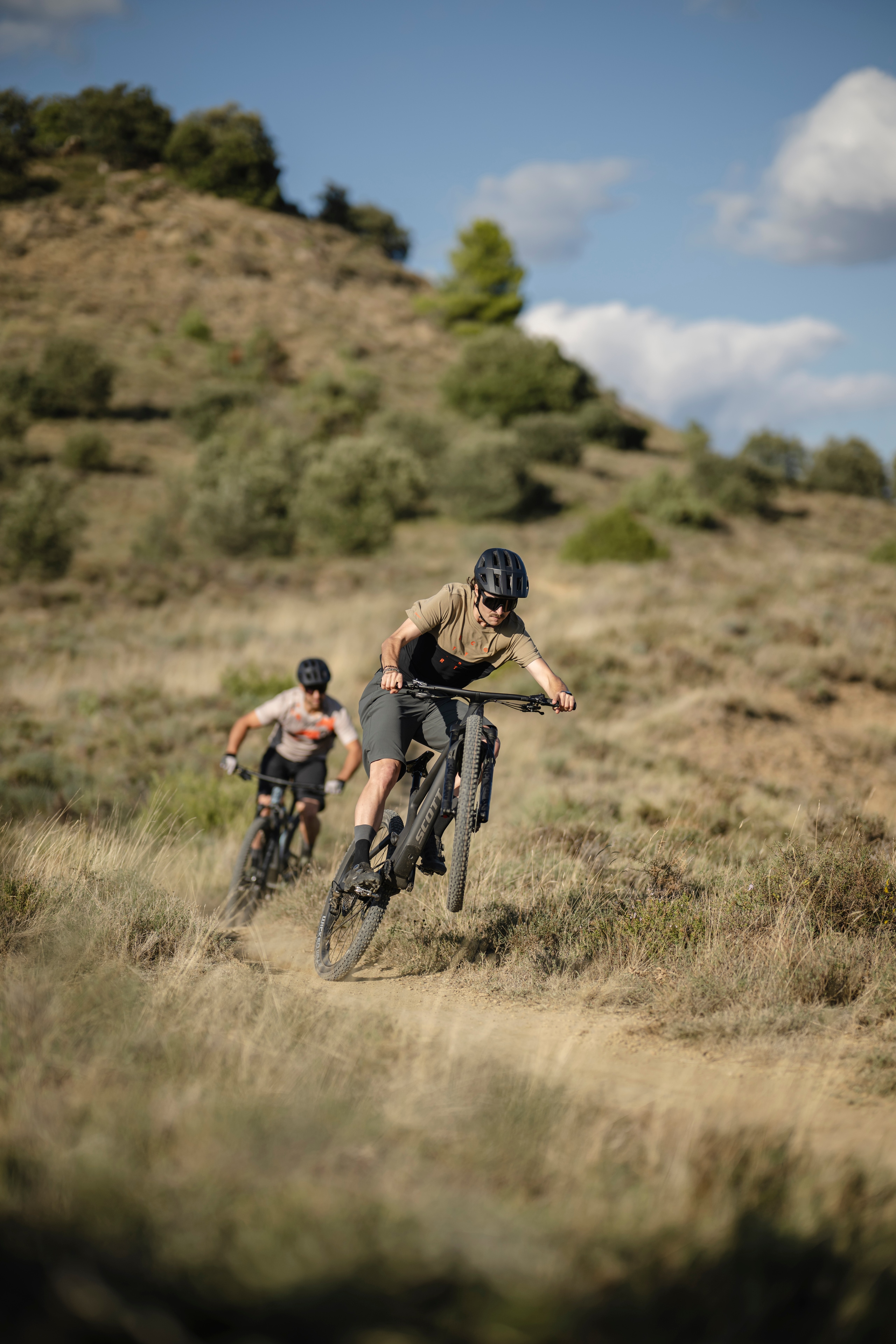 Two people ride Scott mountain bikes through grassy terrain