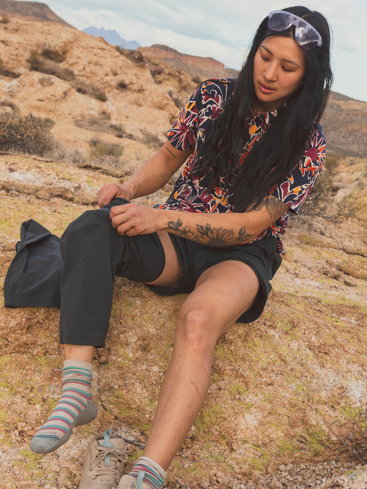 A hiker sits down to zip the legs off of their pants.