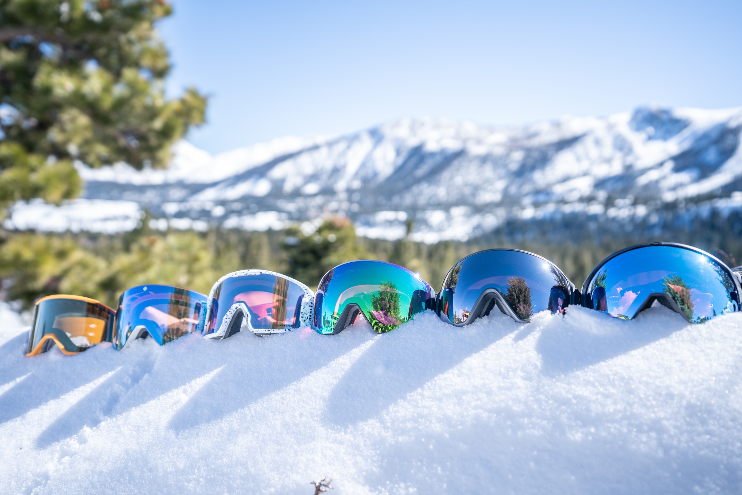 Six goggles are lined up on a snowy ledge against a mountain backdrop.