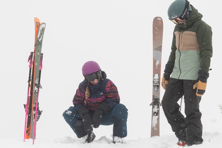 Two skiers stand or squat next to their skis, planted vertical in the snow.