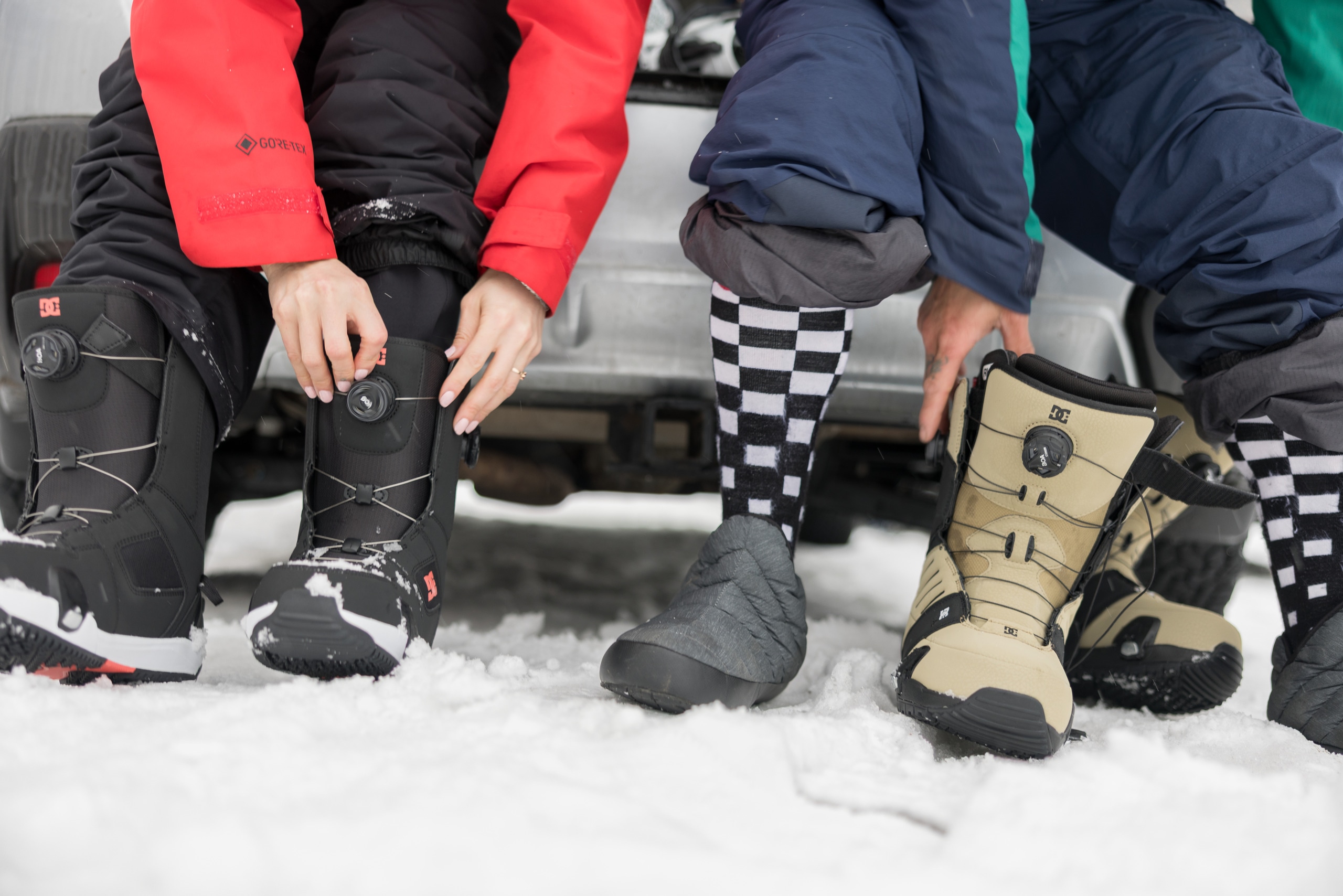 Two snowboarders sit on the back of their vehicle and put on their snowboard boots