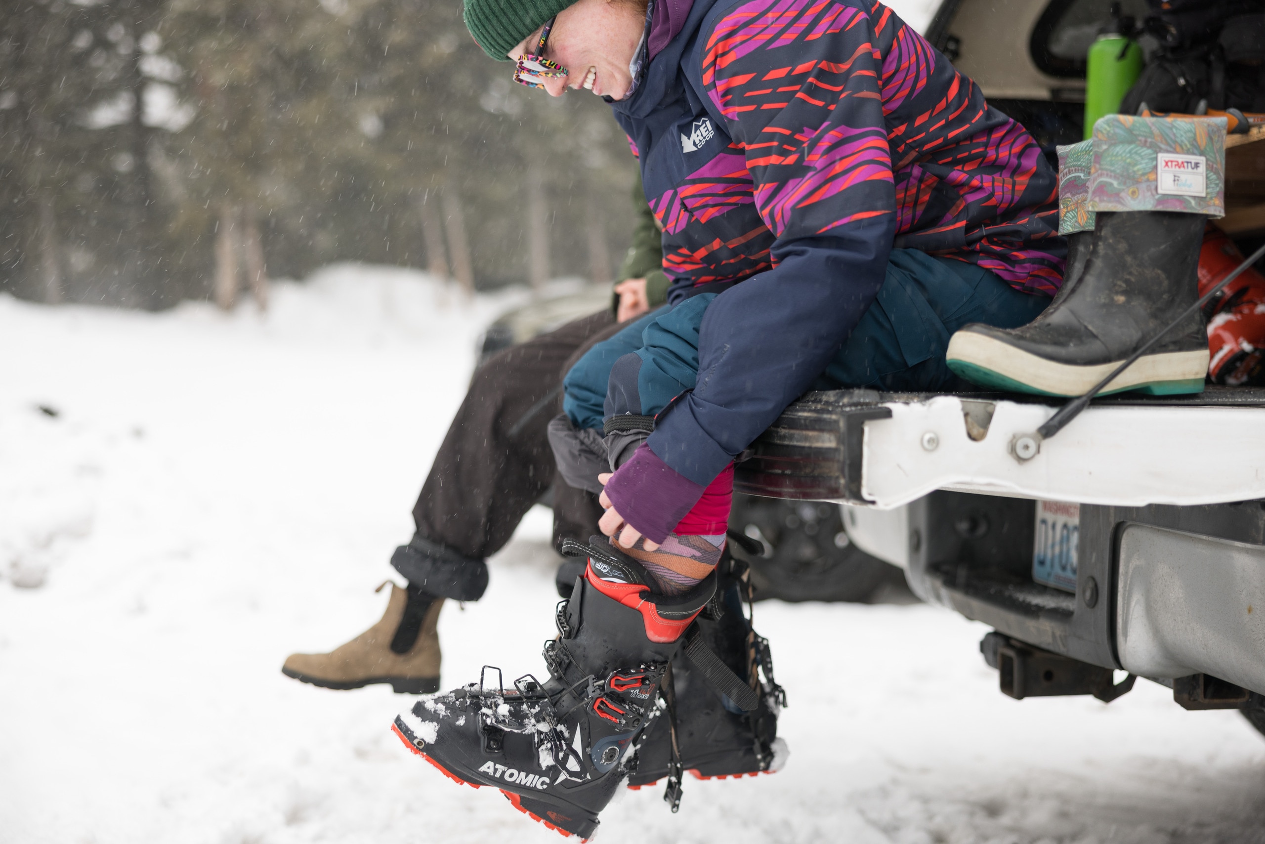 A person sits on the back of a truck to pull on downhill shi boots.
