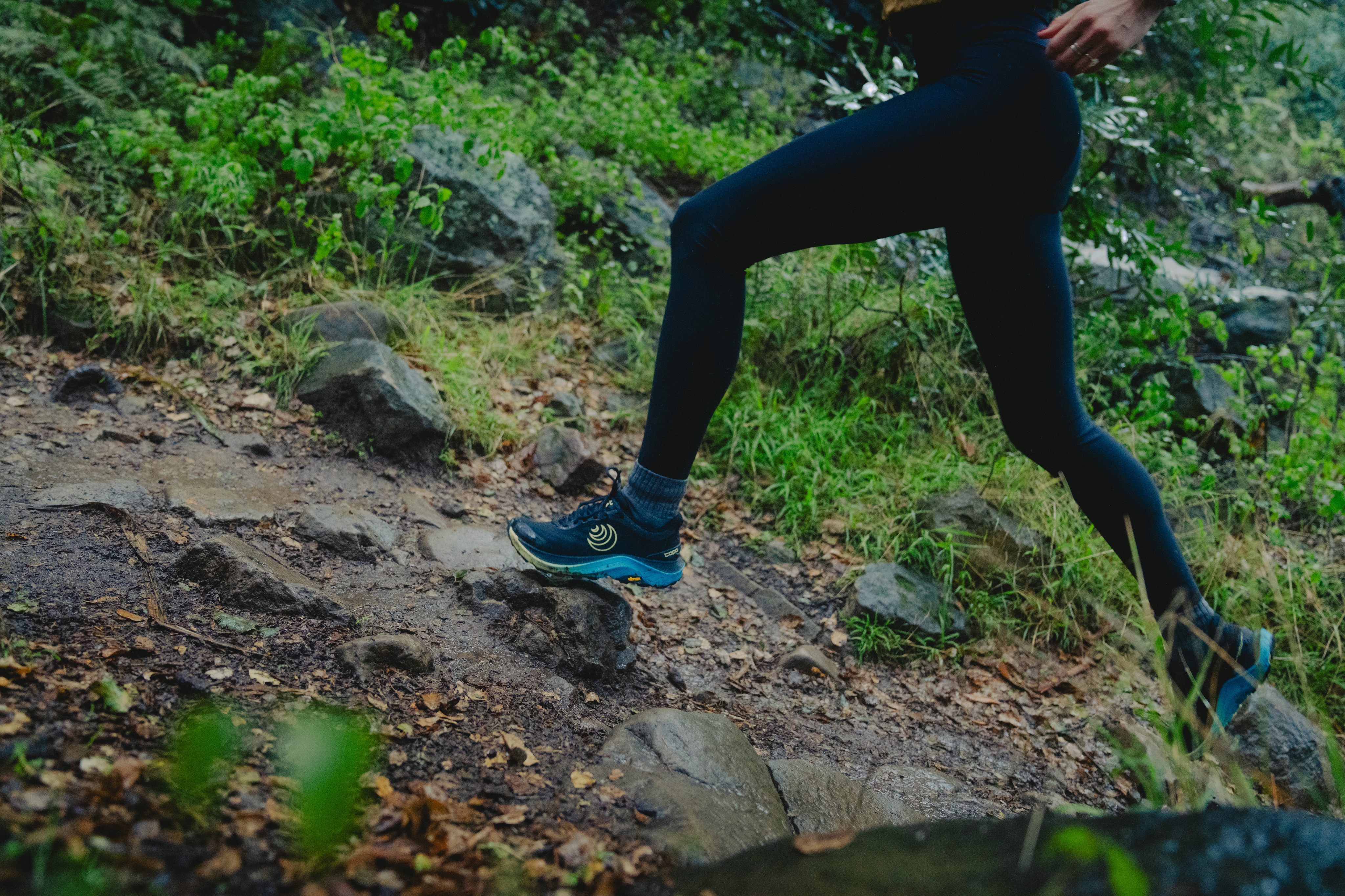 A person wearing Topo Athletic shoes runs through a rocky path on a wet day.