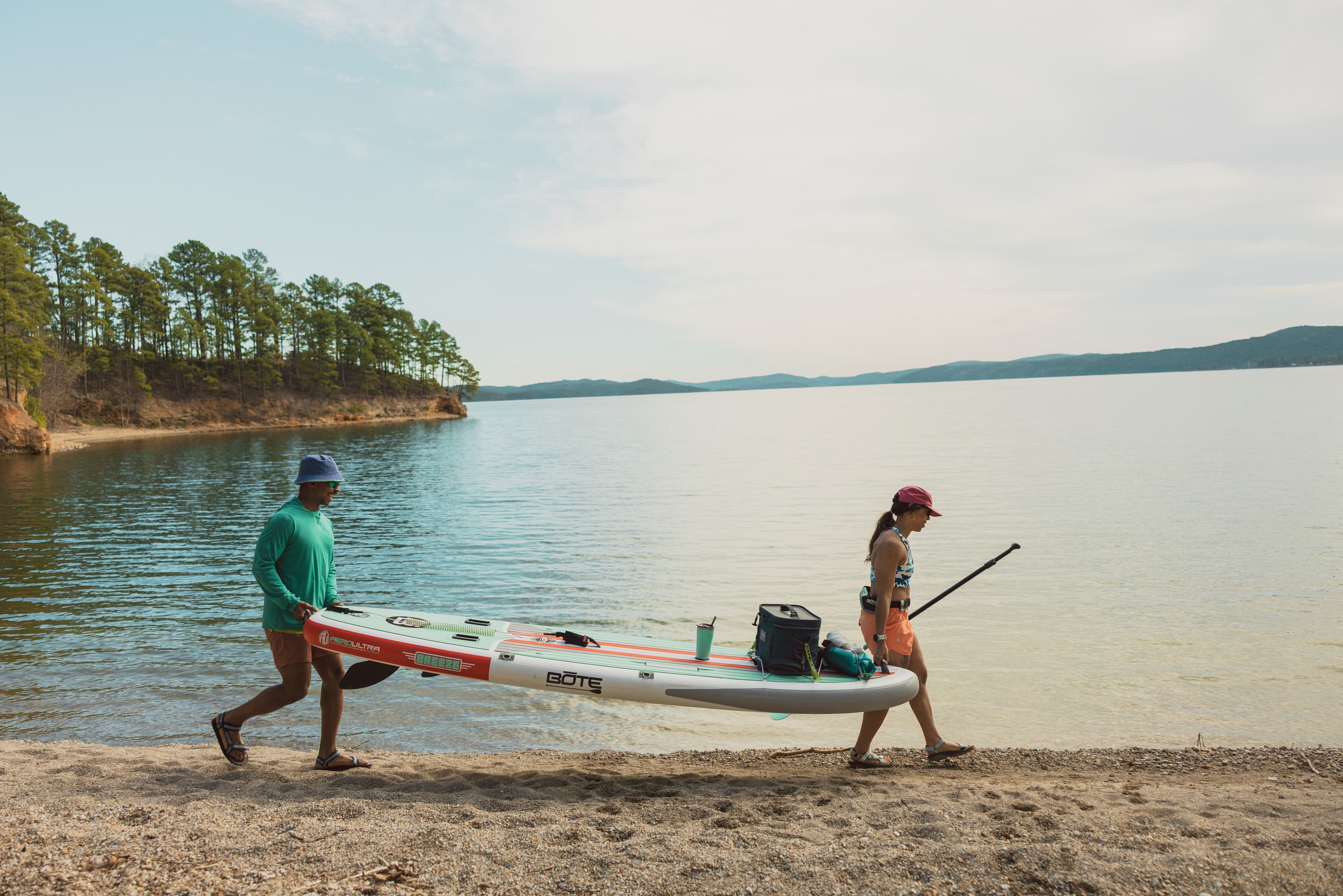 Two people carry a stand up paddle board along a lake