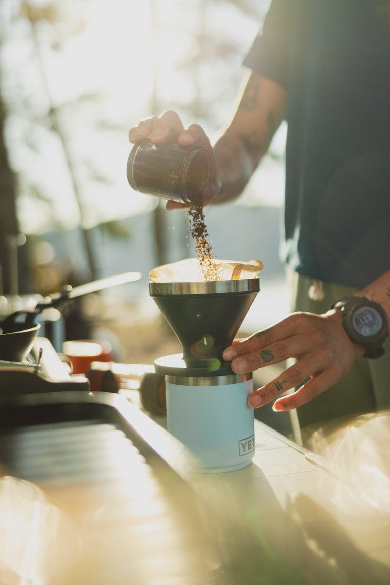 A person pours ground coffee into a pour-over cone at camp