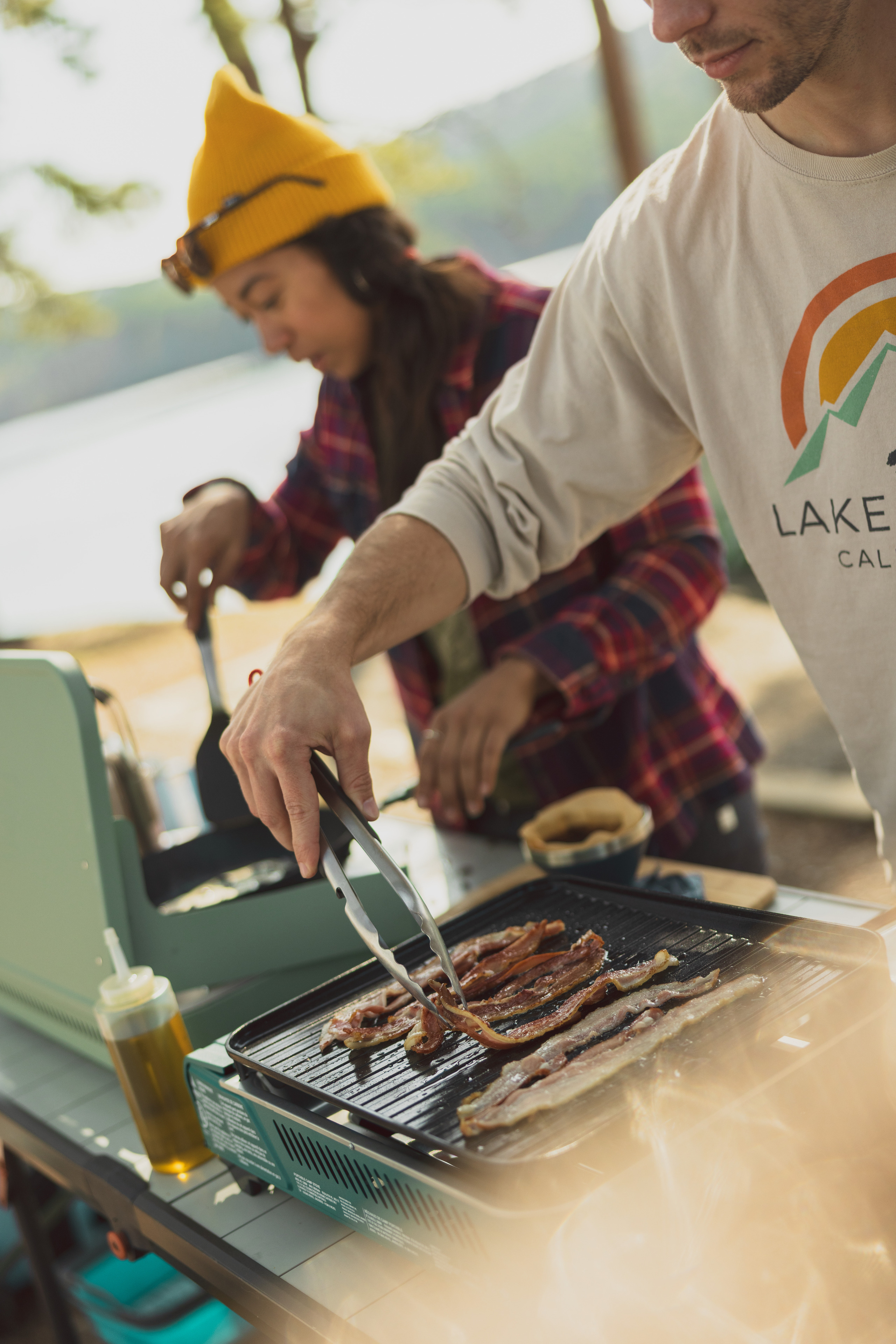 One person cooks bacon on a grill and the other flips pancakes