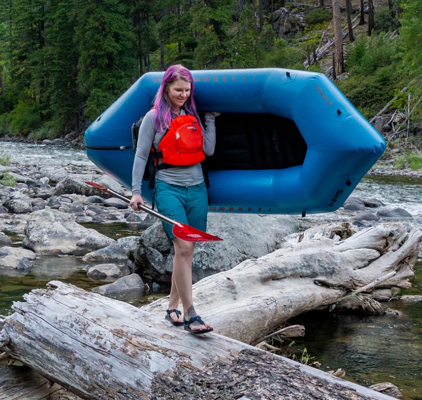 A person walks along a log while carrying a paddle and a packraft.