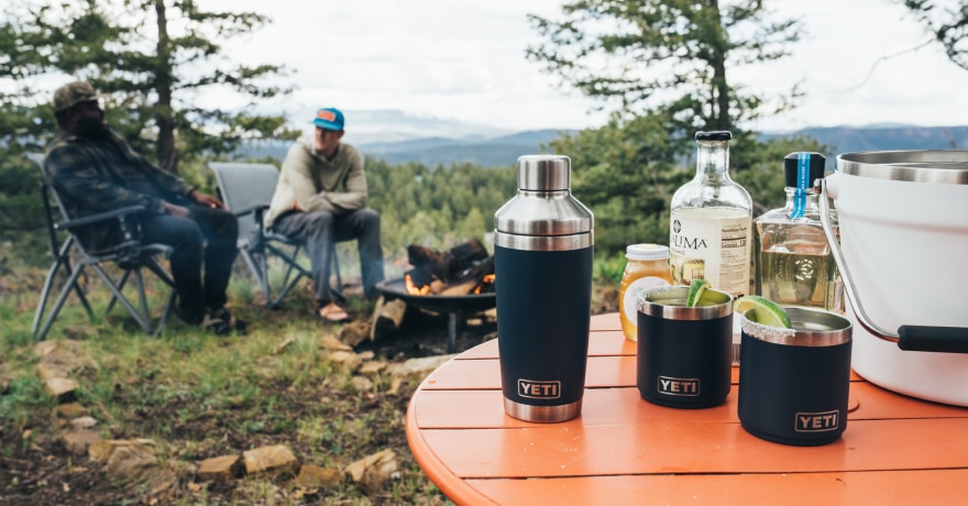 YETI cups, a shaker, and a beverage bucket on top of a table with two people in the background.