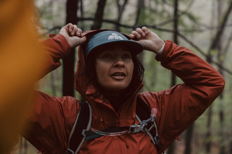 A person pulls their rain jacket's hood up over their head in a wooded area. 