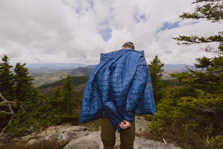 A person on a peak puts on a puffy insulated jacket in front of some pine trees
