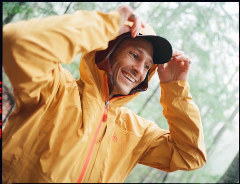 A person wearing a yellow rain jacket in the woods pulls their hood up over their cap.