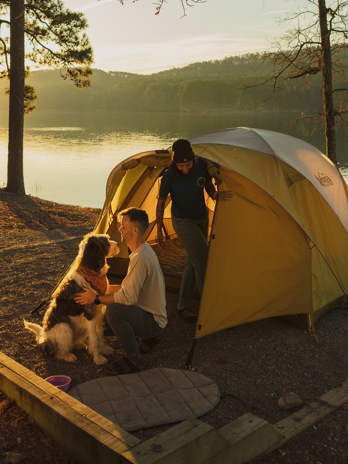 Two people enjoy time with a dog at their campsite.