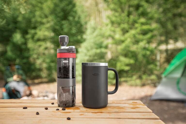 A manual coffee grinder and a coffee mug on a table near a campsite