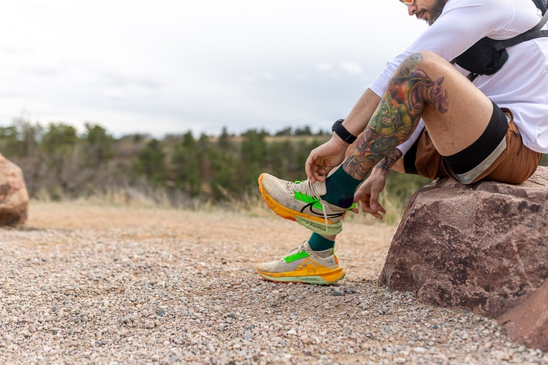 A person sits on a rock and pulls on their trail-running shoes over a pair of crew height socks