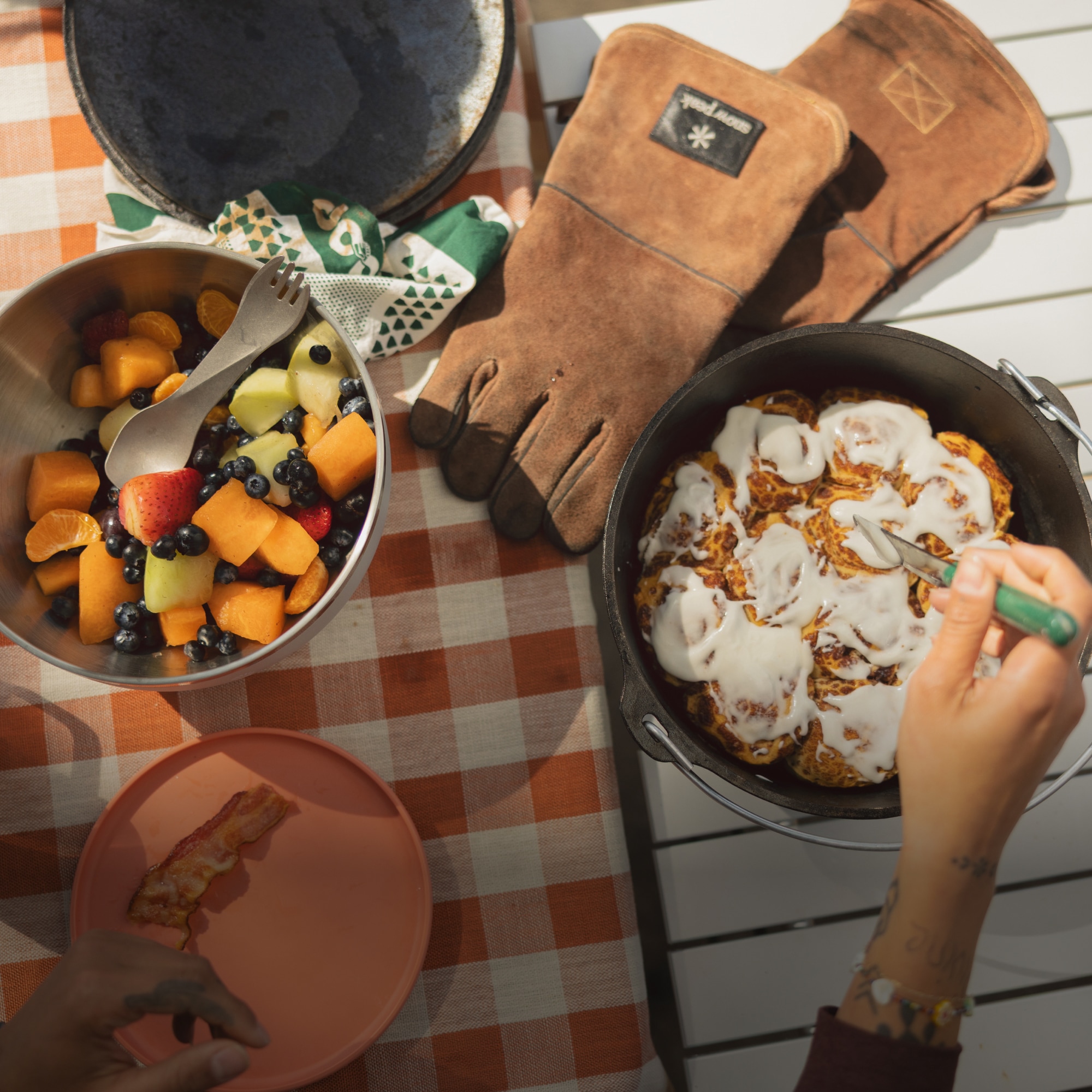 A view of a table from above with a bowl of fruit, cinnamon rolls, a pair of gloves, & plate of bacon.