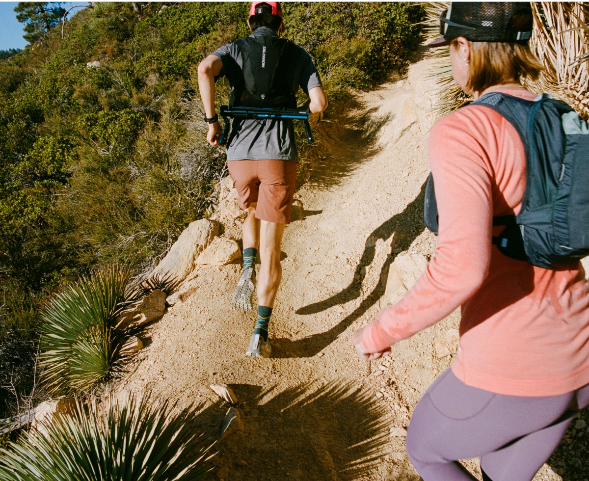 Two people wearing hydration vests run up a rocky trail.