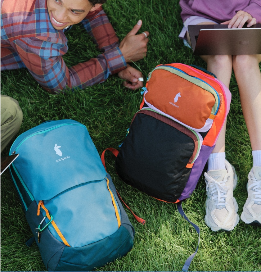 A group of people relax in the grass after class with their Cotopaxi backpacks.