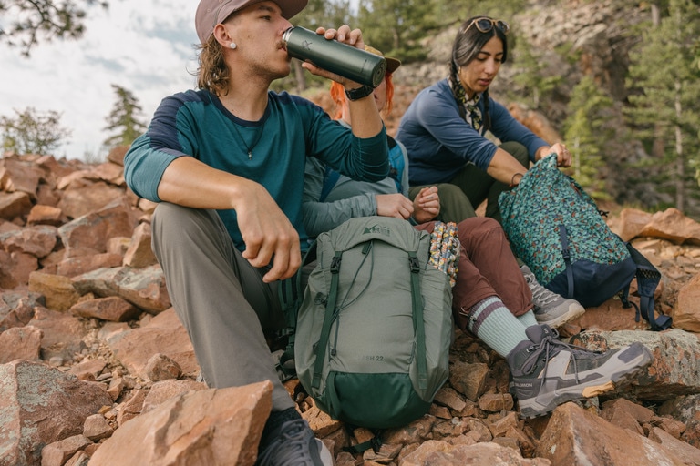 Three people sit on a rocky outcropping with their hiking day packs. One is drinking from an insulated thermos.