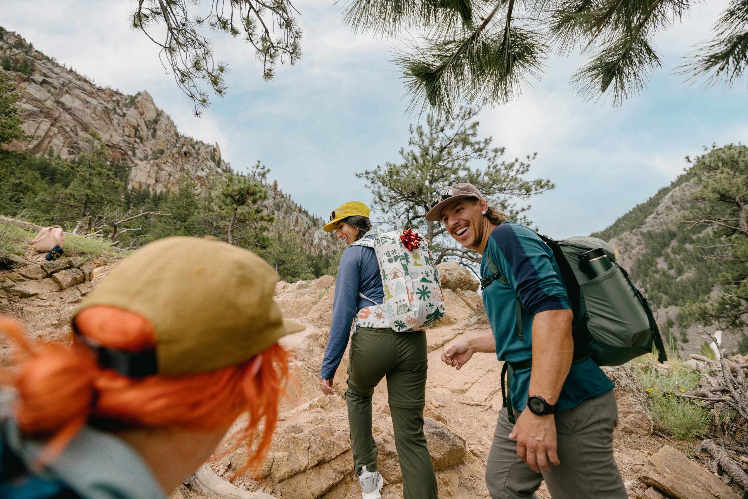 Three people hike up a rocky trail, and the hiker in the lead is wearing a gift-wrapped backpack