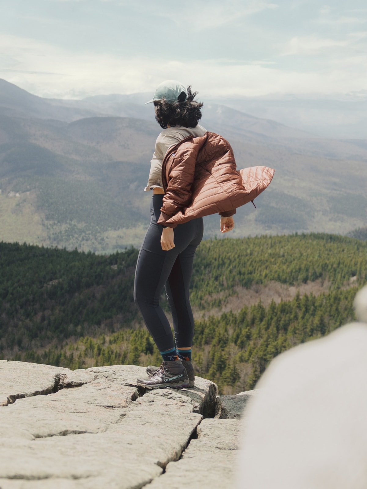 A person stands on a large rock while looking off into the distance.