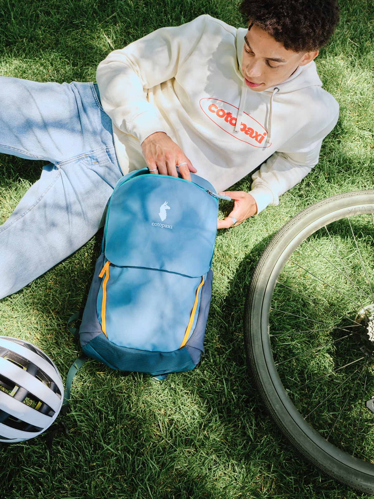 A person relaxes in the grass with their Cotopaxi backpack, a bike, & a bike helmet.